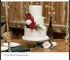 a white wedding cake with red flowers and greenery sits on a table next to plates