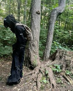 a person wearing a black outfit standing next to a tree trunk in the middle of a forest