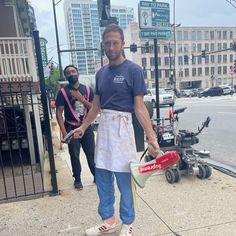 a man in an apron is holding a bucket on the sidewalk while another man stands next to him