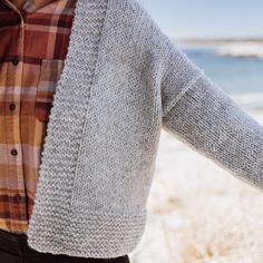 a woman wearing a plaid shirt and cardigan standing on the beach with her hands behind her back