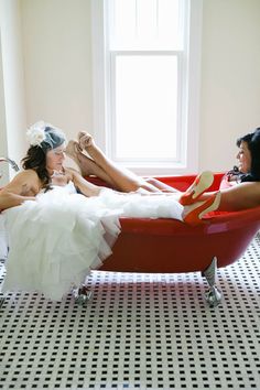 two women in dresses are sitting on a red chair and one is wearing a white dress