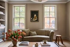 a living room filled with furniture and two large windows next to a wooden table topped with flowers