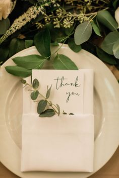 a white plate topped with a napkin covered in greenery and a thank you note