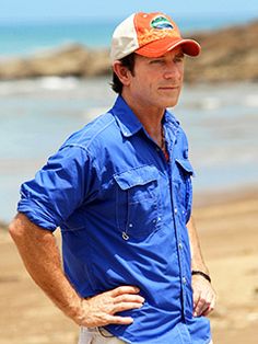 a man standing on top of a sandy beach next to the ocean wearing a blue shirt and orange hat