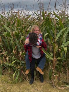 a woman sitting on top of a corn field holding a black dog in her arms