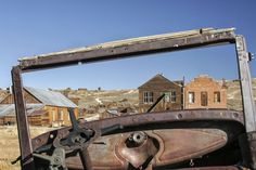 an old rusted out truck sitting in the middle of nowhere with buildings in the background