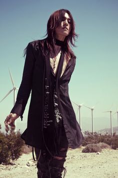 a woman with long hair and black clothes standing in front of wind mills, holding her hand on her hip