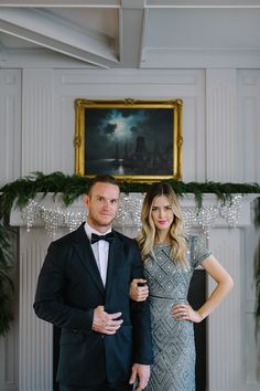 a man and woman standing next to each other in front of a fireplace with christmas decorations