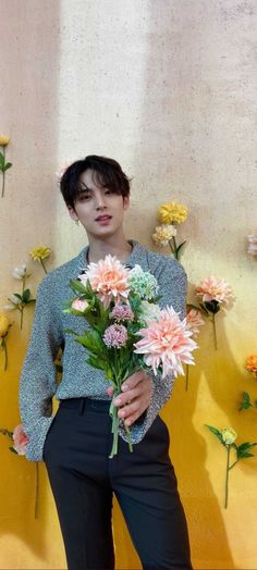 a woman standing in front of a yellow wall with flowers on it and holding a bouquet