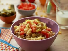 a purple bowl filled with pasta salad next to other bowls of vegetables and milk on a wooden table