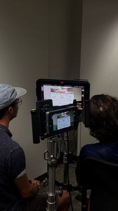 two men sitting in front of a computer monitor on top of a tripod,