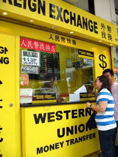 two people standing in front of a yellow foreign exchange kiosk on the street