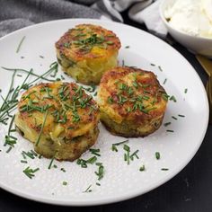 three potato cakes on a white plate next to a bowl of cream cheese and herbs