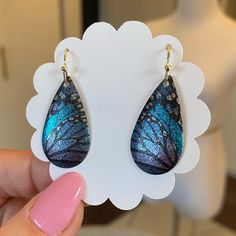 a pair of blue and black butterfly earrings on top of a white card with pink nail polish