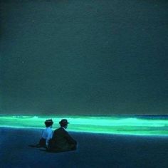 two people are sitting on the beach looking at the green light in the water below