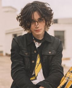 a young man with curly hair sitting in front of a white building wearing a black jacket