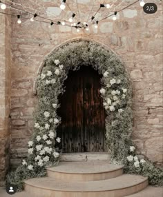 an arch with flowers and greenery on the steps to a stone building, surrounded by string lights