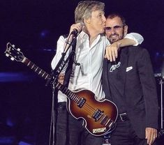 two men standing next to each other while one man is holding a guitar and kissing the other