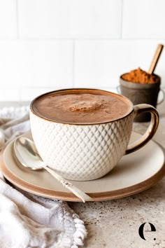 a cup of hot chocolate on a saucer with spoons and cinnamon in the background