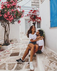 two people sitting on the ground in front of a building with red flowers growing out of it