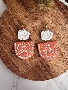 the orange and white flower earrings are sitting on top of a wooden table next to a plant