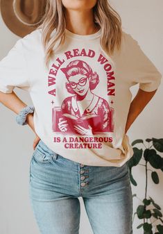 Celebrate the power of knowledge and embrace the spirit of rebellion with this shirt design that boasts a vintage 1950s illustration of a woman immersed in a book. As she captivates the era's charm, the surrounding text boldly declares, "A well-read woman is a dangerous creature." Ideal for book lovers, feminists, and banned book activists, this design encapsulates the strength and influence that come with intellectual prowess.  The juxtaposition of the retro aesthetic with the empowering messag Bookish Crew Neck Shirt With Graphic Print, Relaxed Fit Graphic Print Shirt In Bookish Style, Relaxed Fit Graphic Print Shirt With Bookish Style, Literary Graphic Print Crew Neck T-shirt, Bookish Cotton Shirt With Graphic Print, Literary Cotton Relaxed Fit T-shirt, Literary Style Cotton T-shirt With Relaxed Fit, Bookish Cotton Tops With Character Print, Cotton Slogan T-shirt