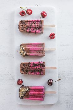 three popsicles with chocolate and cherry toppings on a white plate next to cherries