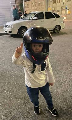 a little boy wearing a helmet and giving the thumbs up sign while standing in front of a car