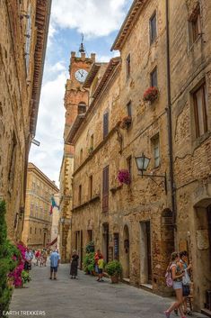 people are walking down an alley way in the old town with stone buildings and flowers