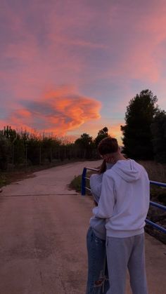 a man and woman standing next to each other on a dirt road under a pink sky