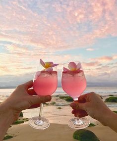 two people holding up wine glasses with pink liquid and flowers on the beach at sunset