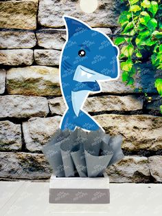 a paper cut out of a blue dolphin on top of some rocks and plants in front of a stone wall