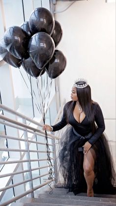 a woman in a black dress is holding some balloons on the stair case while wearing a tiara