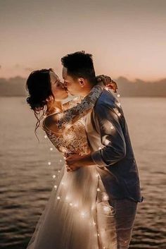 a bride and groom kissing in front of the water at sunset with lights on them