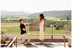 a man kneeling down next to a woman on top of a wooden bench in front of a sign