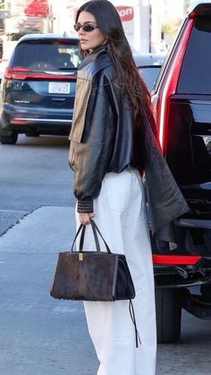 a woman in white pants and black leather jacket carrying a brown handbag while standing next to a red car