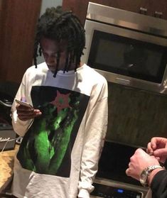 a man with dreadlocks standing in a kitchen looking at his cell phone while another person looks on