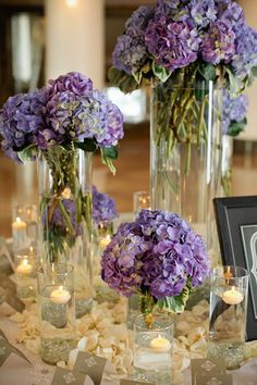 three vases filled with purple flowers sit on a table next to candles and framed pictures