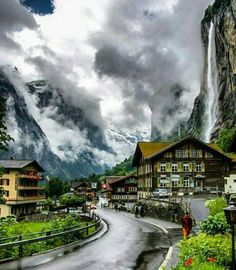 a scenic view of a mountain town with a waterfall in the background and people walking on the road