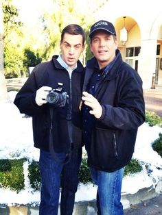 two men standing next to each other in front of snow covered bushes and trees, taking a selfie