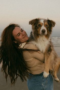 a woman with long hair holding a dog on the beach in front of the ocean