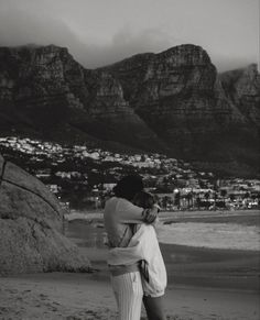 black and white photograph of two people hugging on the beach with mountains in the background