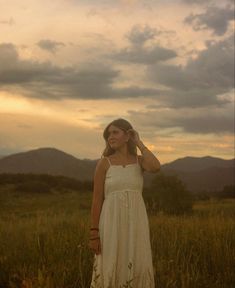 a woman in a white dress standing in tall grass with mountains in the back ground