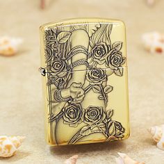 a gold lighter with roses on it sitting on the ground next to shells and seashells