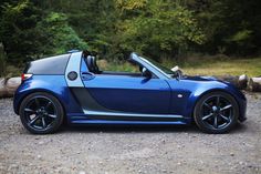 a blue sports car is parked on the gravel near some logs and trees in the background