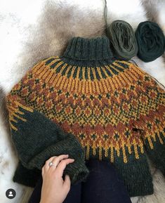 a woman's hand is knitting a sweater on top of a white fur rug