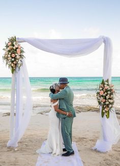 a bride and groom kissing on the beach under an arch decorated with flowers, greenery and ribbons