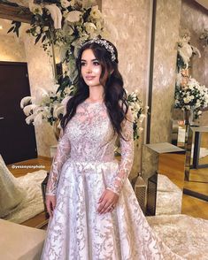 a woman standing in front of a mirror wearing a wedding dress with long sleeves and an elaborate headpiece