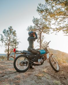 a man on a motorcycle talking on the phone while standing next to some pine trees