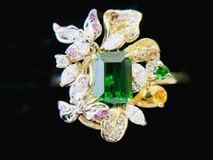 Introducing our exquisite forest green Tsavorite ring, adorned with natural diamonds, colored sapphires, and rubies. This one-of-a-kind statement piece is sure to turn heads with its vivid color and intricate details. Inspired by the delicate beauty of flower petals, this ring has been expertly handcrafted with great attention to detail. The intense green color of the Tsavorite gemstone is perfectly complemented by the bi-color gold band, creating a stunning burst of color that will easily match Luxury Tsavorite Jewelry With Gemstone Accents, Green Flower-shaped Gemstone Jewelry, Green Multi-stone Ruby Ring, Fine Jewelry, Luxury Tsavorite Multi-stone Emerald Ring, Colored Sapphires, Green Multi-stone Tsavorite Jewelry, Tsavorite Ring, Delicate Beauty, Be Natural